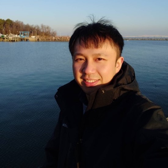 A smiling color photo of Xin Gu wearing a winter coat outside in front of a body of water.