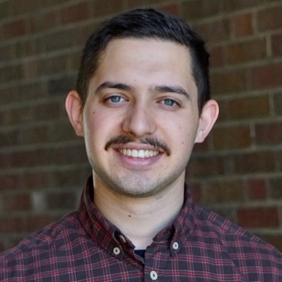 A smiling photo of Ilir Isufi with dark hair, wearing a red plaid shirt with a brick wall in the background.