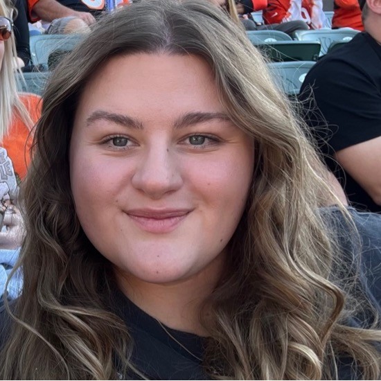 A smiling photo of Hannah Dahlke with wavy brown hair, seated in a stadium with green seats and people in the background.