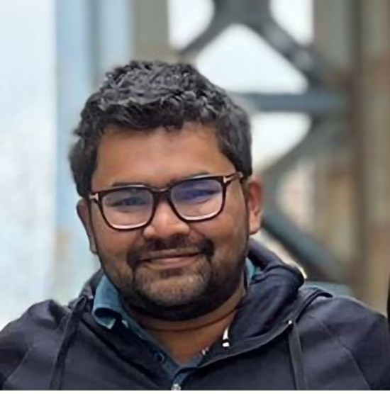A smiling photo of Asif Rayhan with dark hair, wearing glasses and a dark jacket, with blurry architecture in the background.