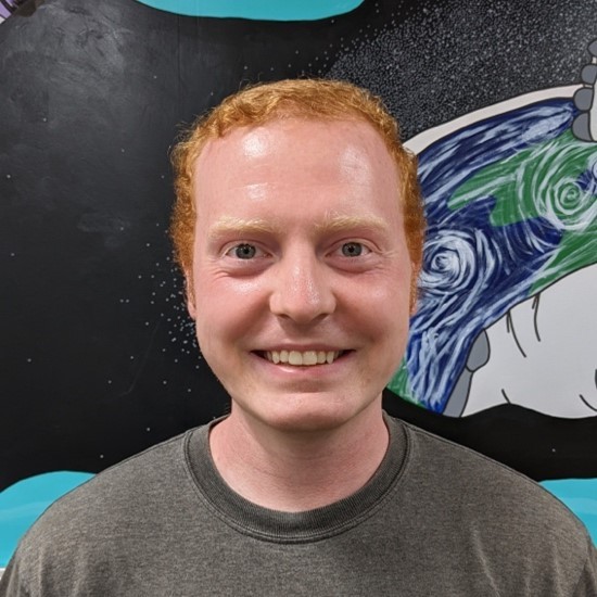 A smiling color photo of Alex Olivan wearing a grey shirt in front of a painted wall.