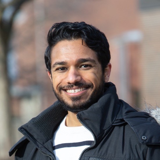 A smiling photo of Sanjay Prasher with dark hair, wearing a dark jacket with blurry architecture and trees in the background.