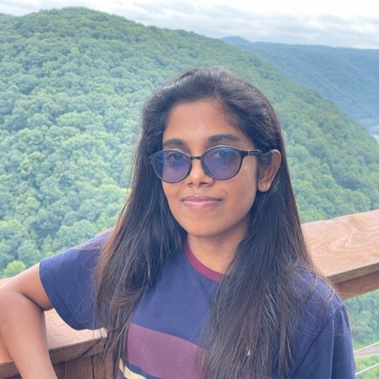 A smiling photo of Tharushi Rajaguru with dark hair, wearing sunglasses and a striped colored shirt, leaning against a rail, with rolling, forrested mountains in the background.