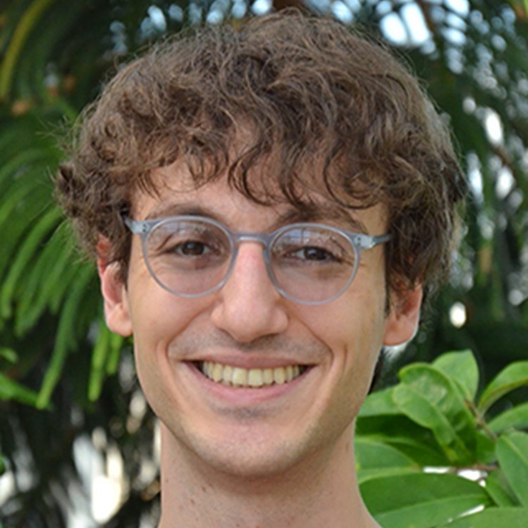 A smiling color photo of Deniz Korman wearing glasses in front of a leafy background.