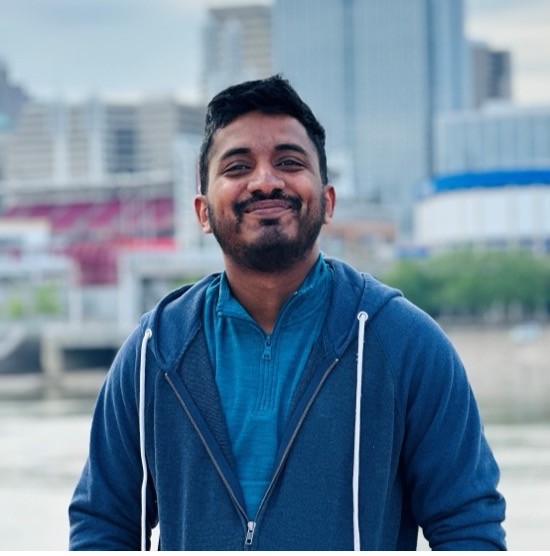 A smiling photo of Thiraj Dhanushan Mohankumar with dark hair, wearing a blue shirt and sweatshirt, with a city skyline and trees in the background.