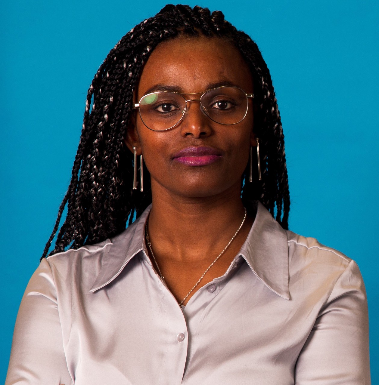 A color photograph of Abeba Birhane wearing glasses, earrings, and a necklace with a silver collared shirt in front of a blue background.