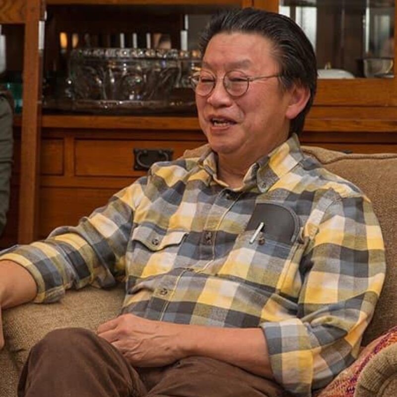 A color photograph of Abeba Birhane wearing glasses, earrings, and a necklace with a silver collared shirt in front of a blue background.