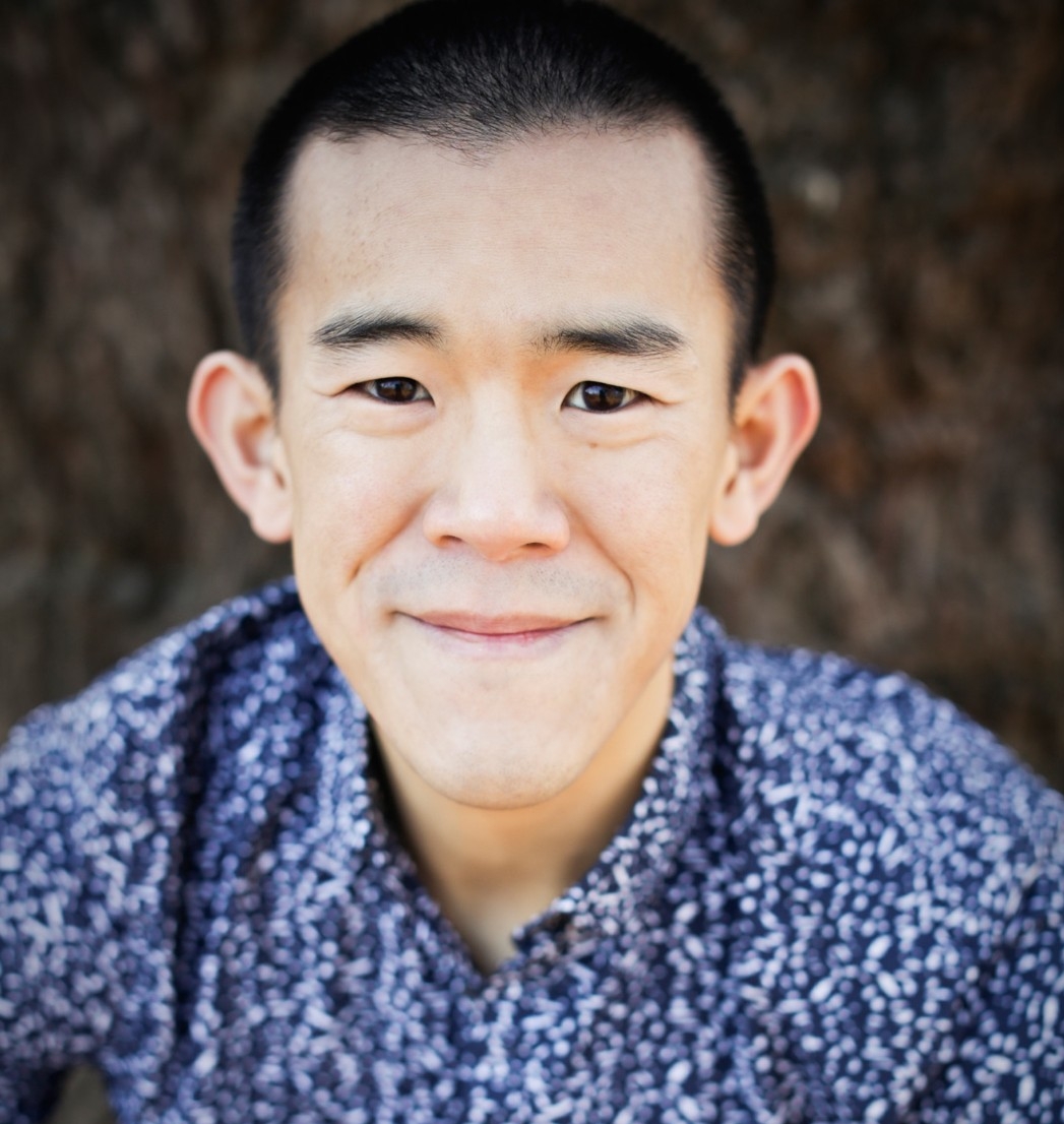 A color photograph of Abeba Birhane wearing glasses, earrings, and a necklace with a silver collared shirt in front of a blue background.