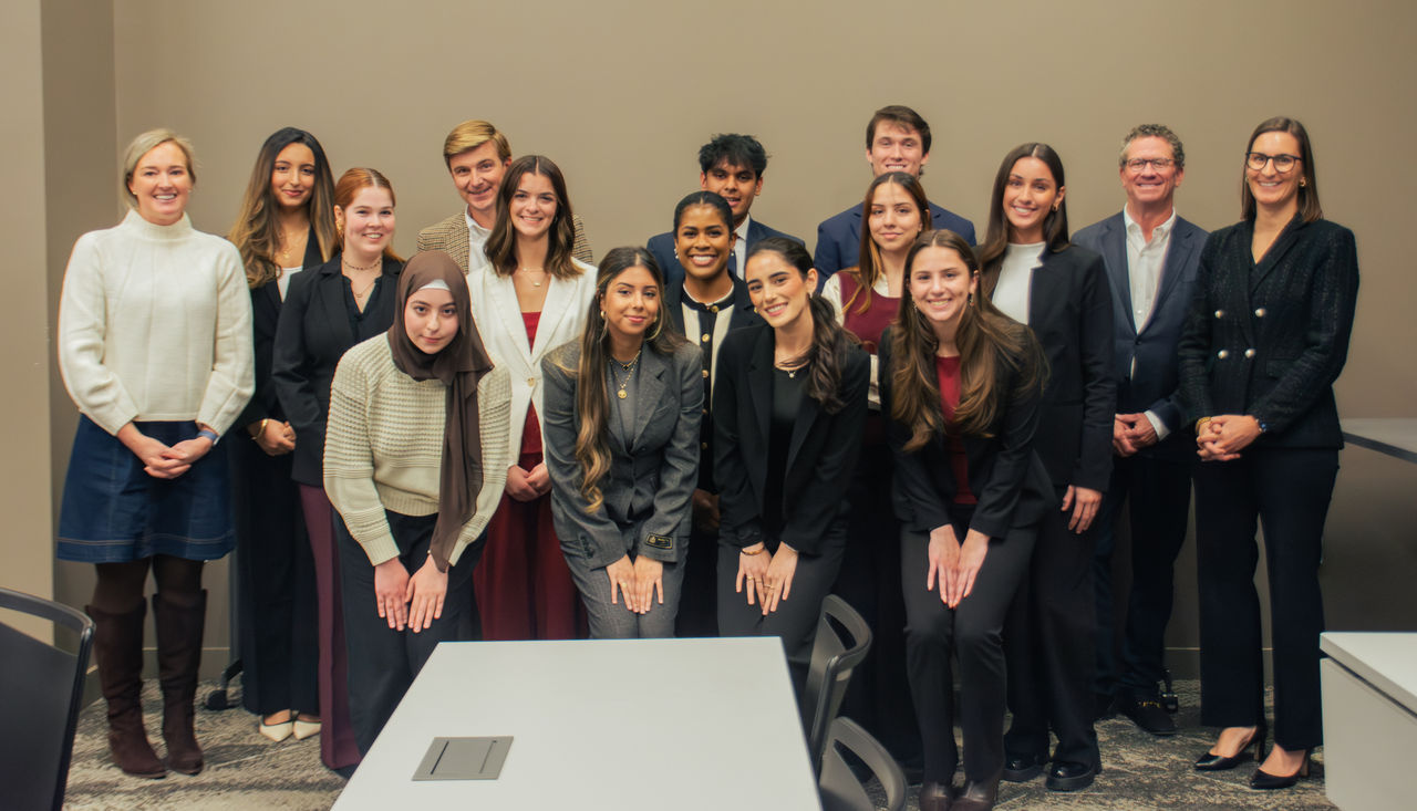 A diverse group of 15 people, including men and women, pose and smile in business attire. The setting is a formal indoor office environment, conveying professionalism.