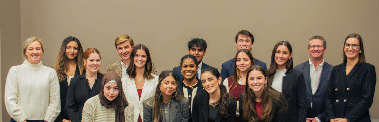 A diverse group of 15 people, including men and women, pose and smile in business attire. The setting is a formal indoor office environment, conveying professionalism.