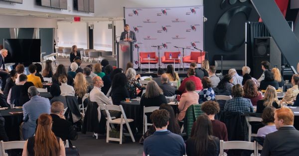 At the end of a long indoor event space, a man stands behind a podium and addresses an audience.