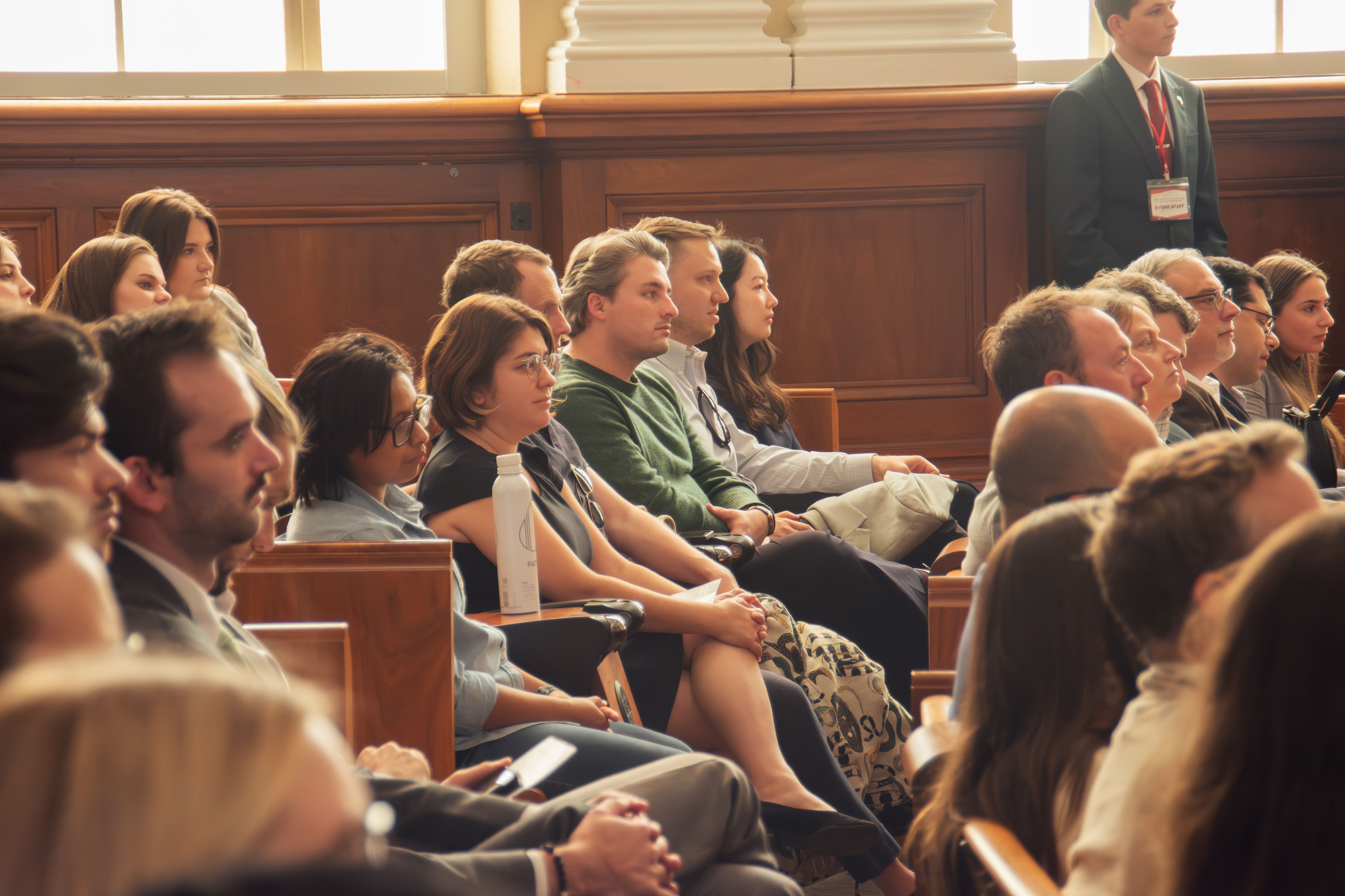 Audience attentively listens at a conference in a wood-paneled room. People of various ages and ethnicities are seated, absorbing the presentation.