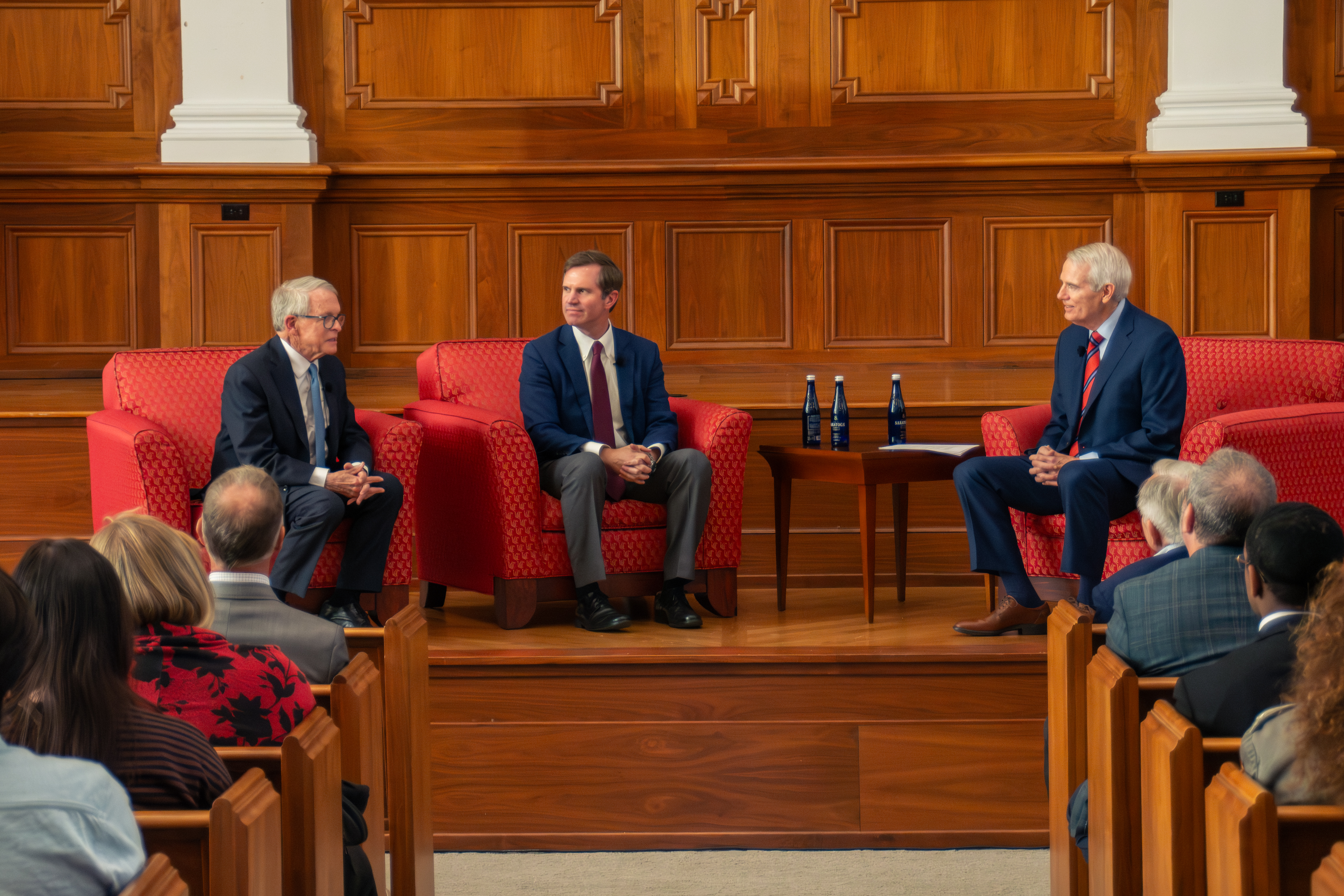 Three men in suits sit on red armchairs discussing in a wood-paneled room. An attentive audience is visible in the foreground. The atmosphere is formal.