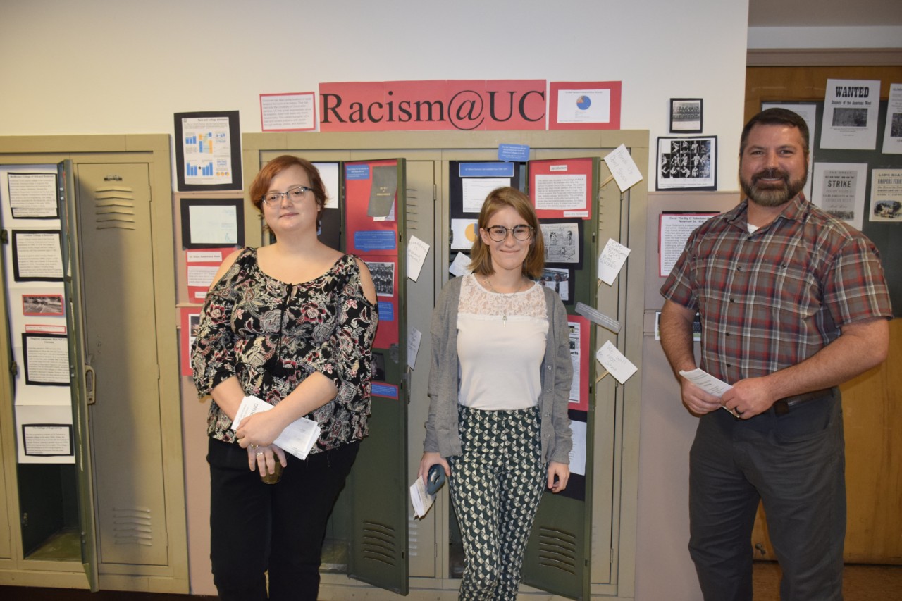 Three students standing in front of locker exhibits with title "Racism @ UC"